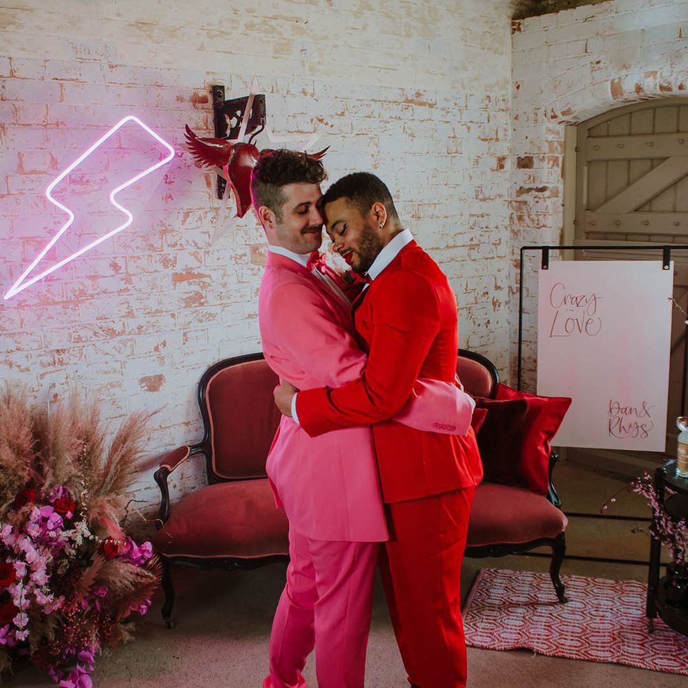 Groom in a red suit embracing his partner in a pink suit next to a pink neon lightning bolt sign and a velvet sofa 