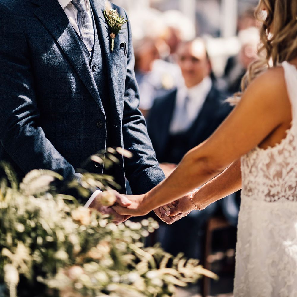 Groom in three piece blue suit with spotted tie smiles at the bride as he holds her hands during the ceremony 