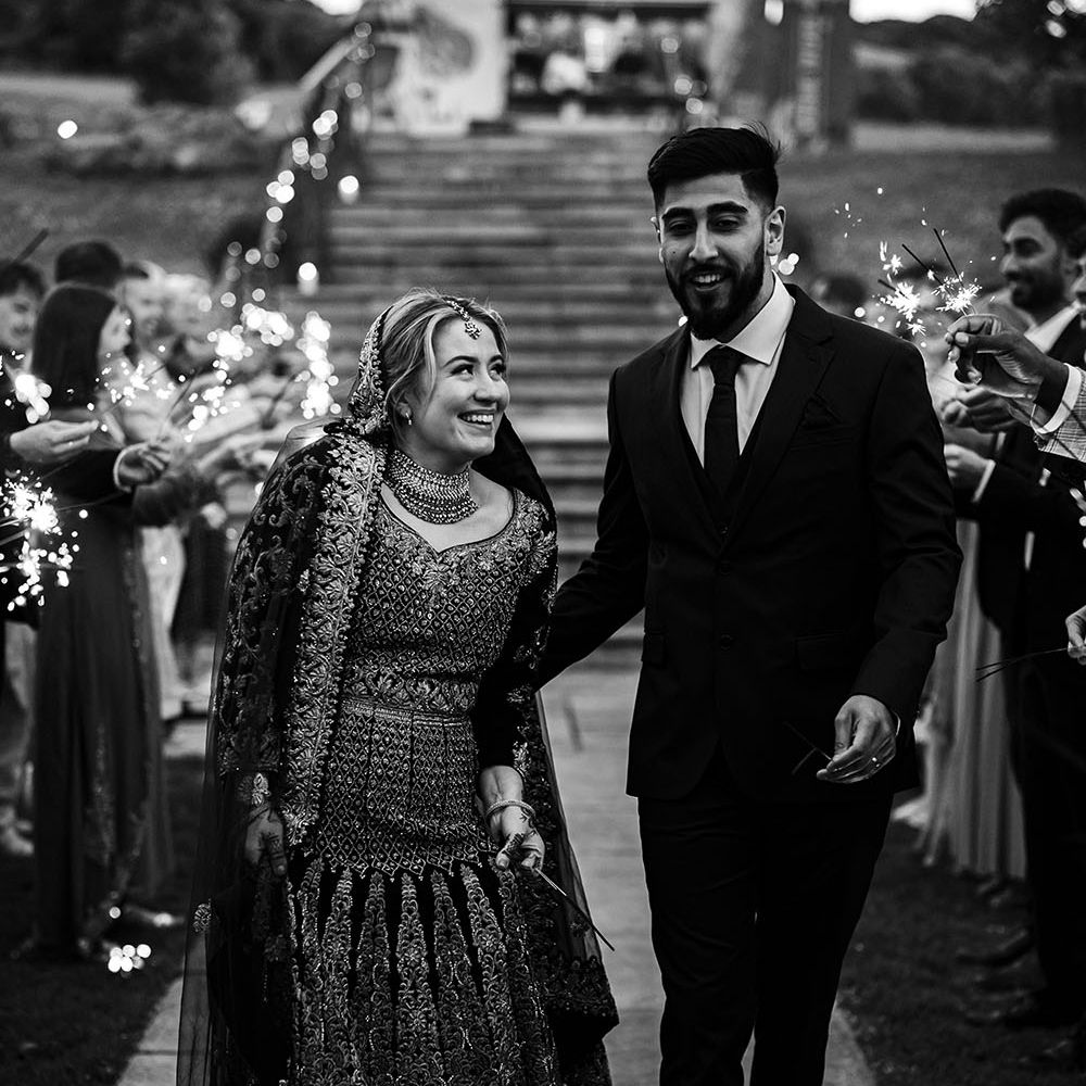 Bride looks lovingly at her groom as they walk through outdoor sparkler exit in black and white image