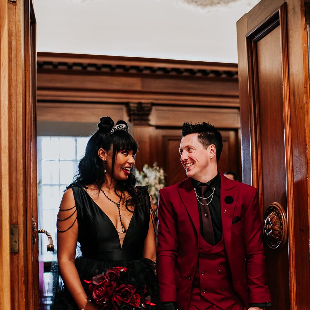 Bride wears black wedding dress and walks with her groom in red suit complete with black shirt 