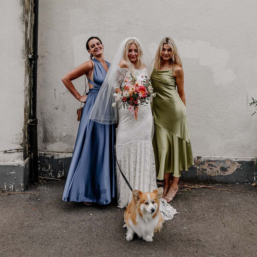Bride in boho two piece lace bridal outfit with large colourful mixed bouquet standing with two bridesmaids in mismatched satin dresses