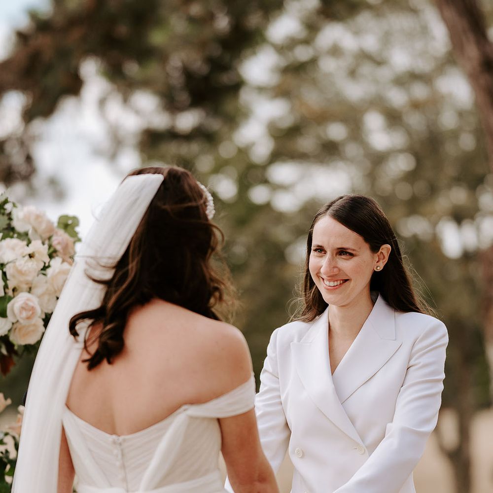 Bride in a white double breasted jacket stands at the altar holding hands with the bride for their outdoor ceremony 