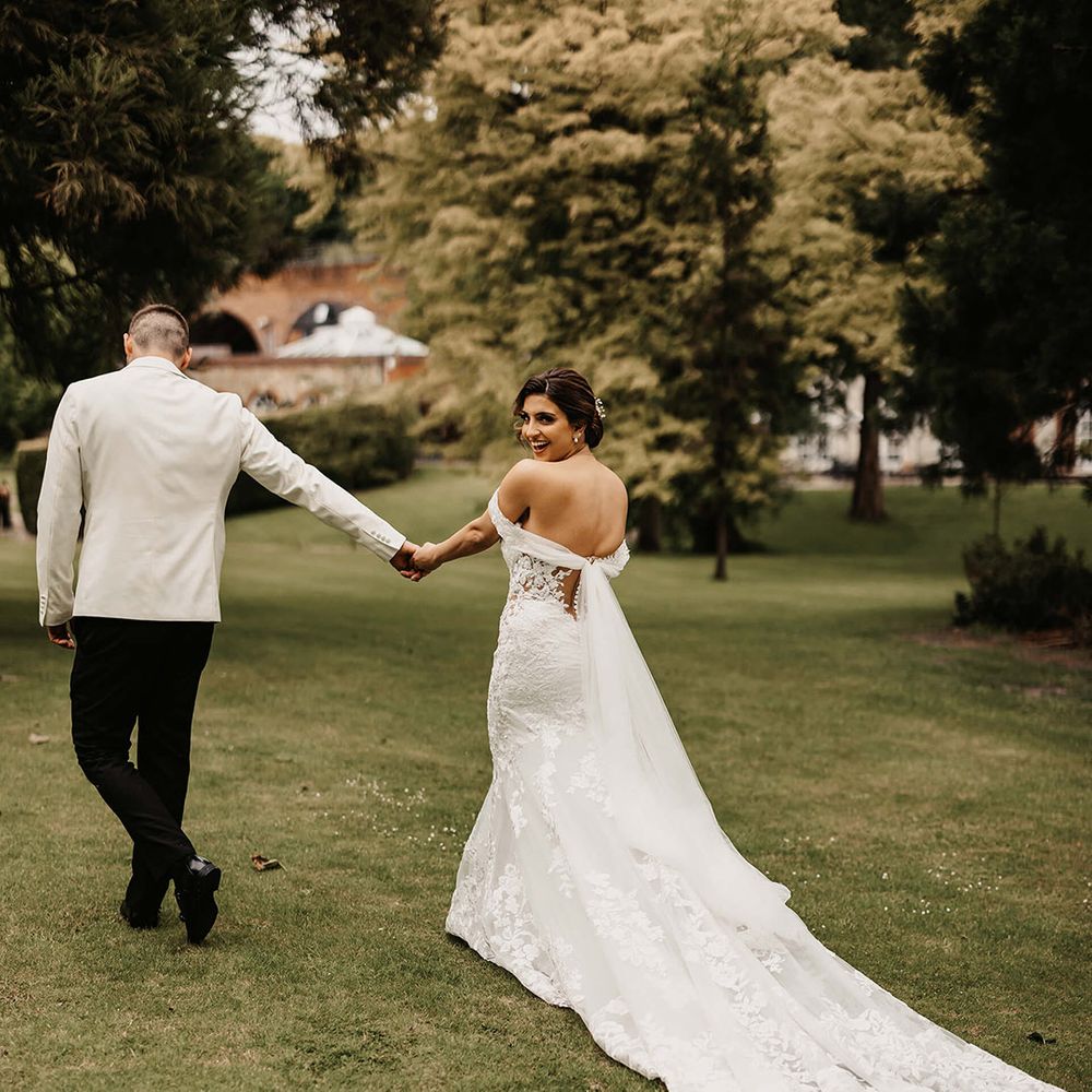 Groom in a white suit jacket walks hand in hand with the bride in an off the shoulder lace wedding dress 