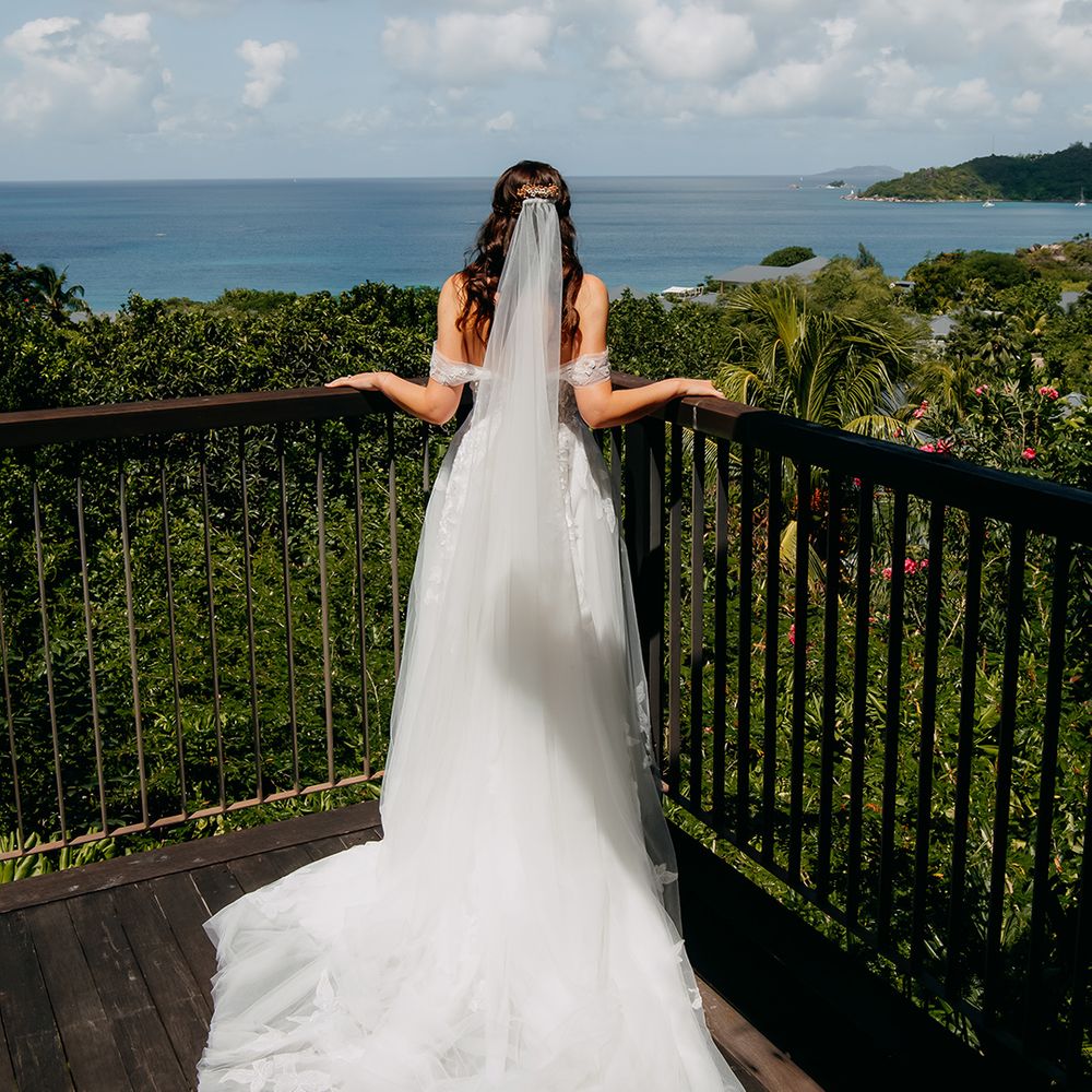 Bride in off the shoulder lace wedding dress with lace floral details and cathedral length veil looking out at Seychelles 