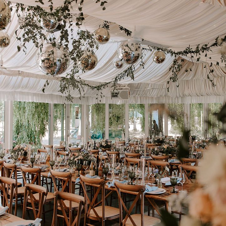 Marquee reception decorated with foliage and disco balls with simple table setting on long wooden banquet tables 
