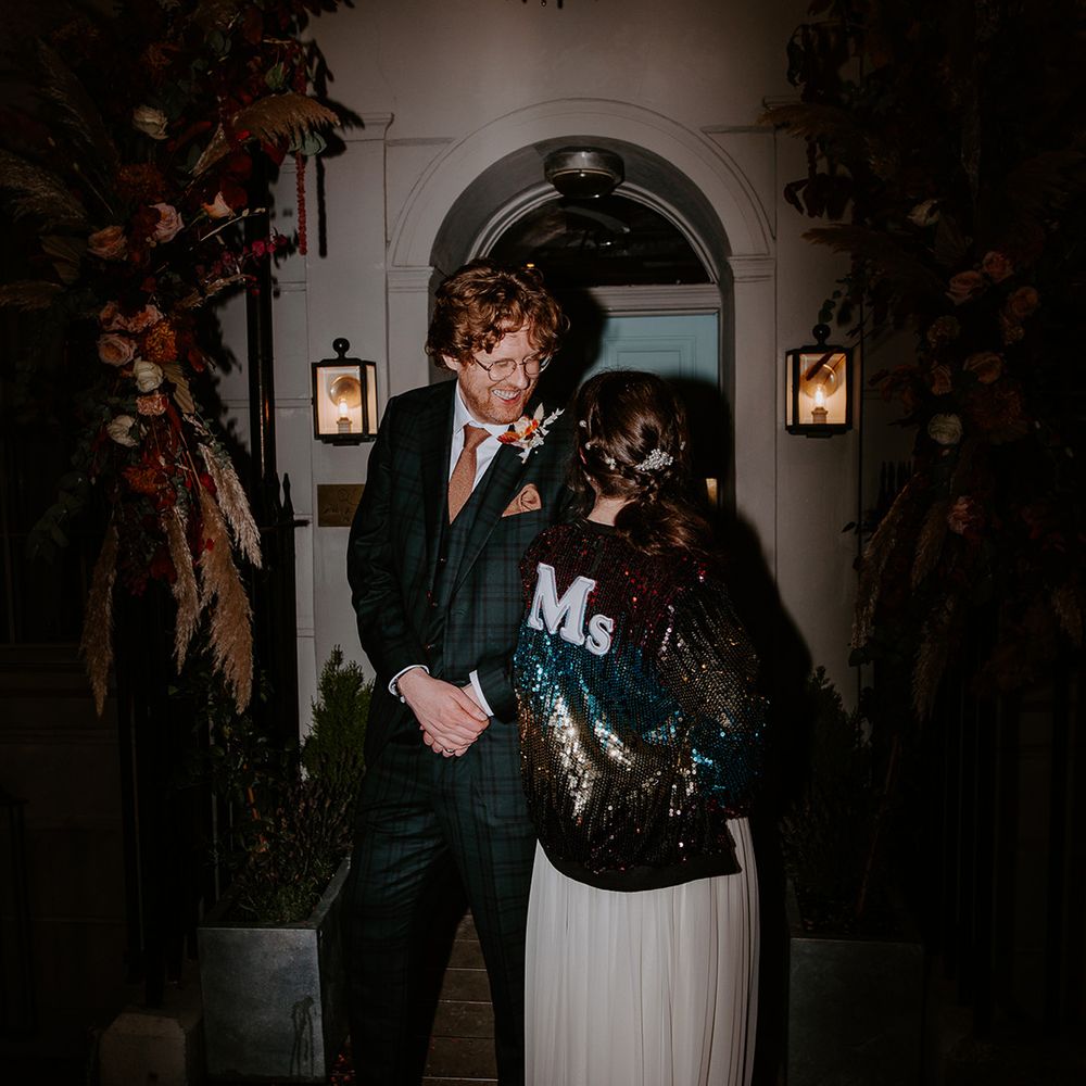 Bride in long vintage wedding dress with sparkly personalised multicoloured wedding jacket looking at groom in dark suit with orange tie at wedding party 