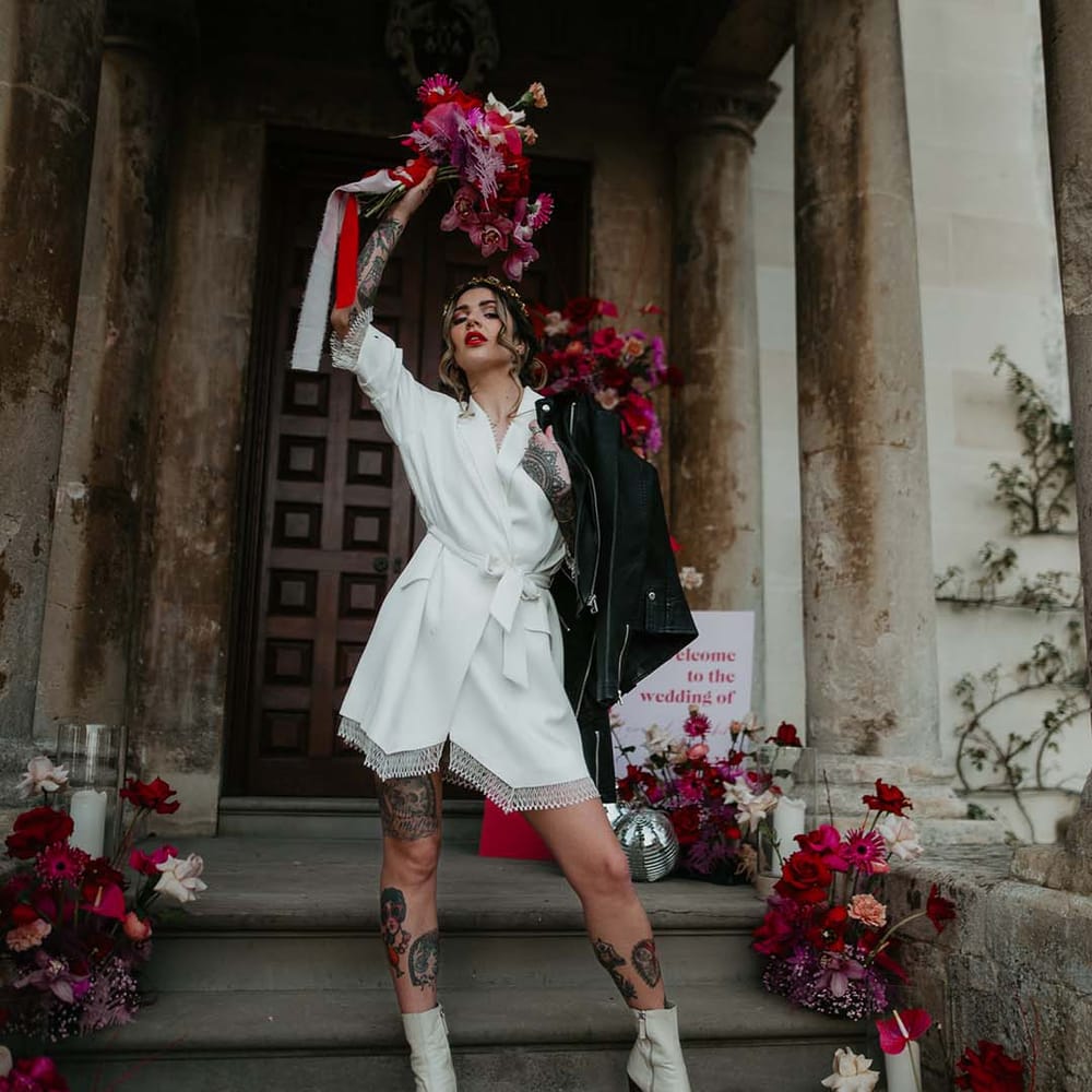 Bride wearing blazer style wrap around short wedding dress with delicate fringe detailing holding personalised, embroidered black leather jacket and wearing short wedding veil with delicate flower embroidery at the base
