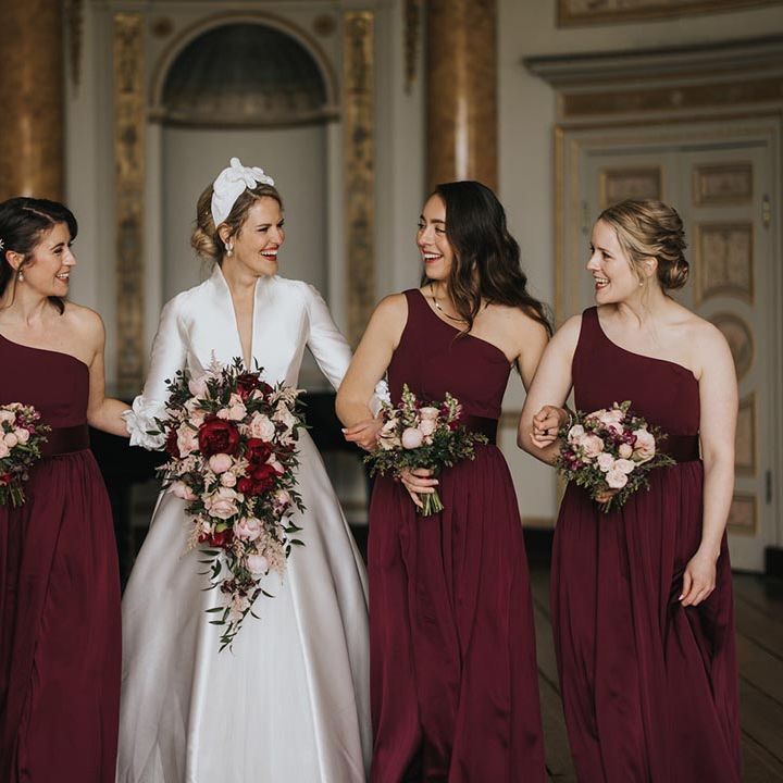 Bridesmaids in burgundy one shoulder dresses smiling with the bride in a conservative high neck long sleeve wedding dress at Stowe House