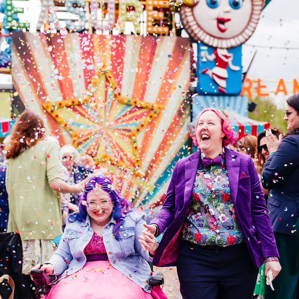Bride in wheelchair wearing personalised denim jacket, pink wedding dress and blue tights holding hands with bride in purple velvet blazer and black suit trousers with purple bowtie, pink patterned pocket square and colourful patterned shirt walking through their colourful confetti throw 