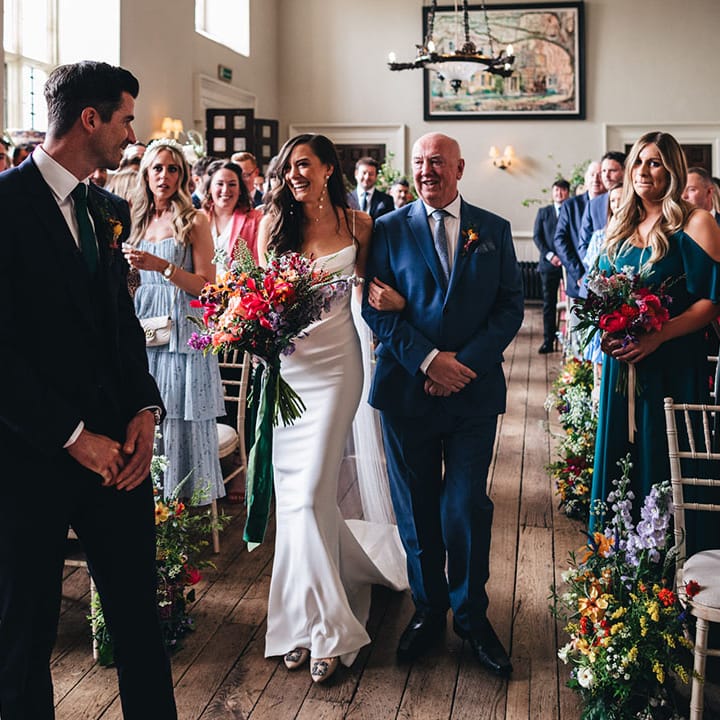 Father of the bride in a blue suit walks the bride to the end of the aisle where the groom waits 
