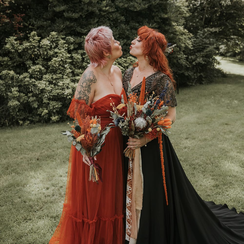 Bridesmaid in burnt orange off the shoulder bridesmaid dress and bride in black dress for wedding in Devon, both holding large dried flower wedding bouquets 