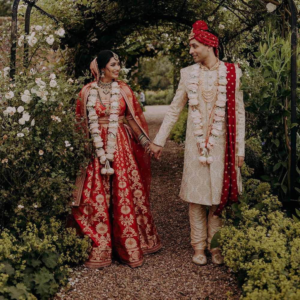 Bride in red and gold lehenga and groom in red, white and gold sherwani stand holding hands in lush gardens 