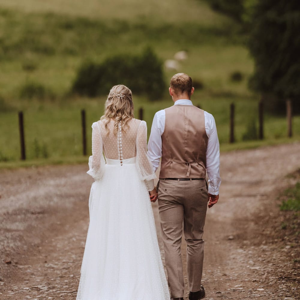 The bride in a Sassi Holford wedding dress walks hand in hand with the groom in a beige waistcoat and trousers 