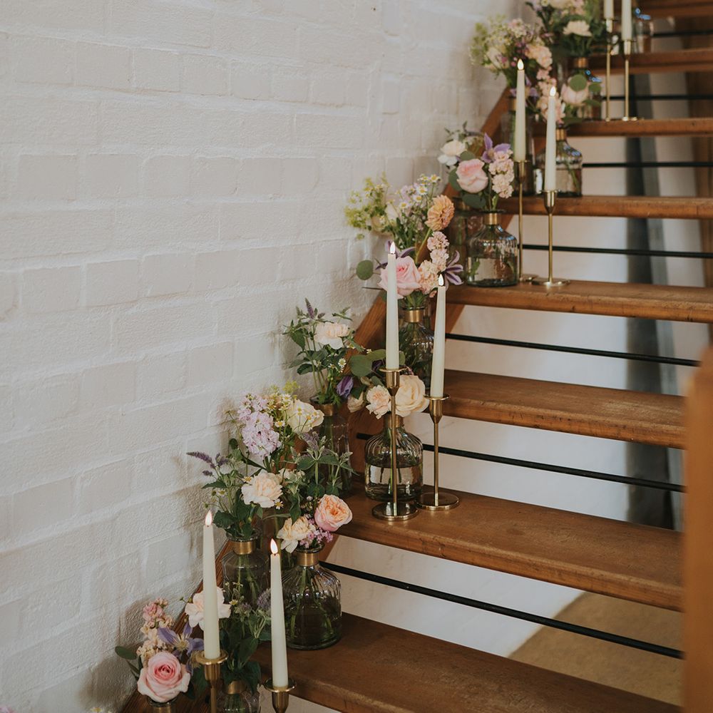 Pastel wedding flowers in shades of pink, purple, cream, and orange with white taper candles in gold holders decorating the stairs 