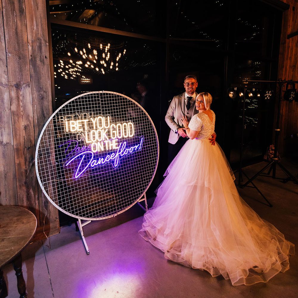 The bride and groom pose next to a decorative neon wedding sign with white and purple neon colours