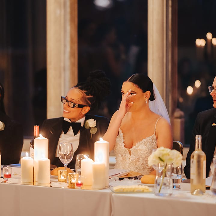Bride wearing lace sleeveless v neck Enzoani wedding dress wiping tear from her eye sitting next to bride in black snakeskin print tuxedo with black bowtie and white rose boutonniere at head table of Grittleton House
