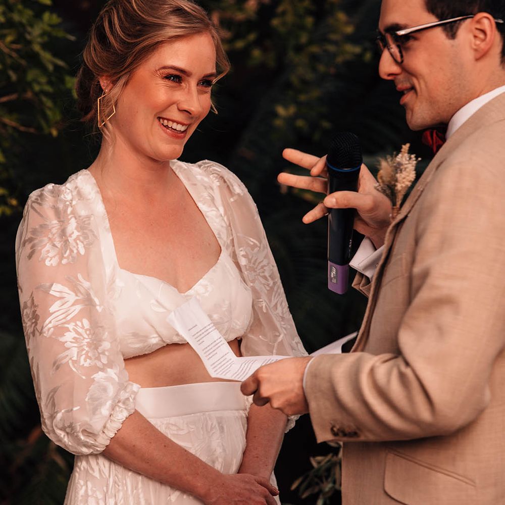 Groom in cream suit reads out his personalised wedding vows with the bride in lace separates smiling brightly at the groom with gold earrings 