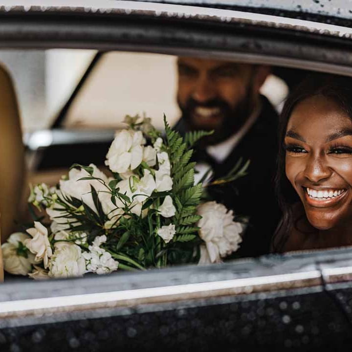 Bride holding white garden rose, white hydrangea, cows parsley and foliage bridal bouquet in classic black wedding car on her way to Lains Barn wedding reception