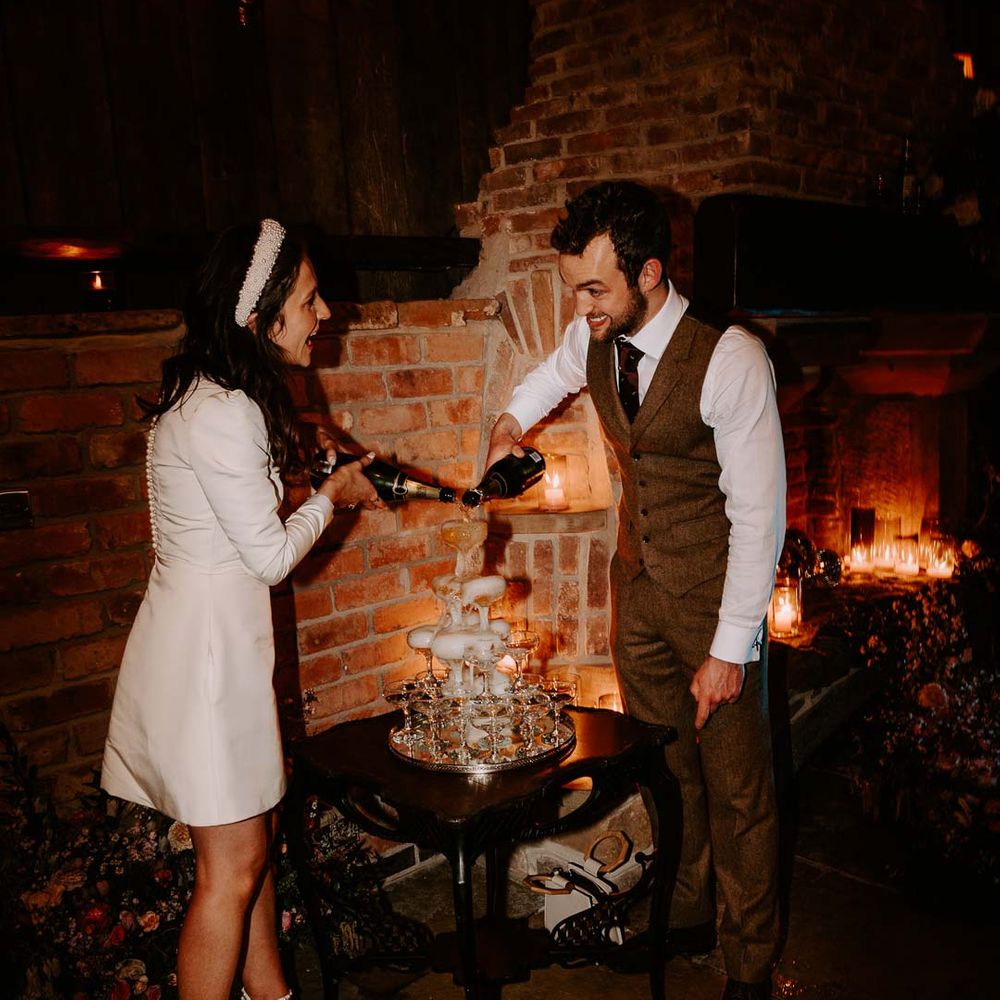 Bride in long sleeve short wedding dress, pearl bridal headband and platform pearl wedding heels standing with groom in brown waistcoat and suit trousers doing wedding champagne tower at Willow Marsh Farm Loughborough
