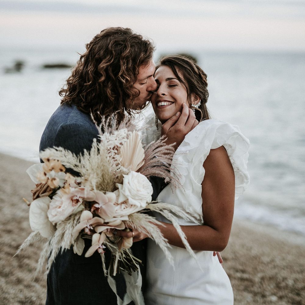 Groom with curly hair kissing his brides cheek on the beach whilst she holds her orchid wedding bouquet with anthuriums and pampas grass