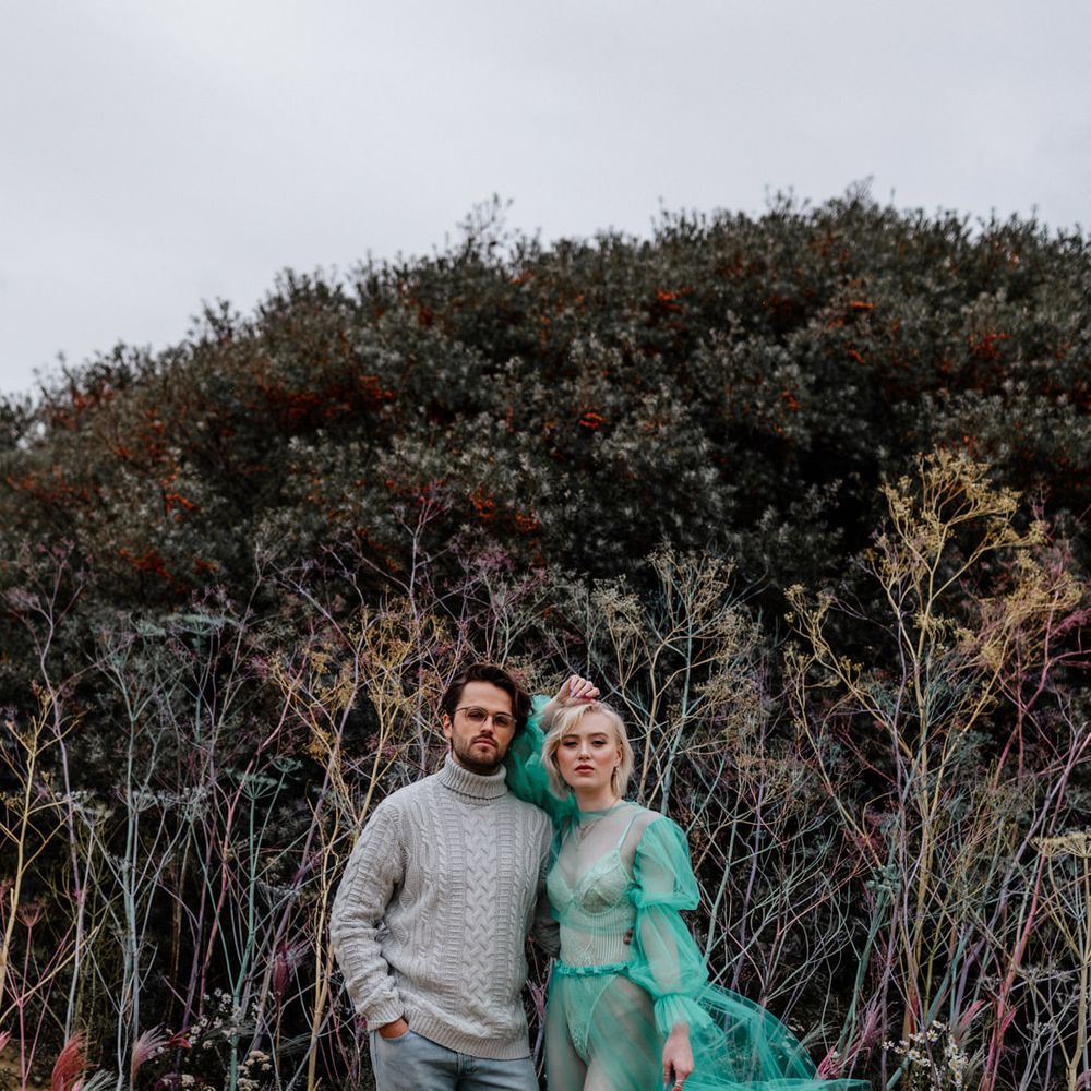 Stylish bride in an all in one body suit with a sheer pastel wedding dress standing neck to her groom on the beach in a grey jumper and jeans 