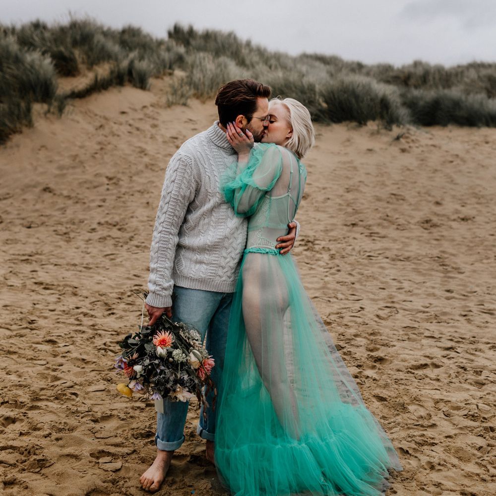 White bearded groom in a grey jumper and rolled up stone wash jeans kissing his bride in a sheer pastel wedding dress on the beach at their elopement 