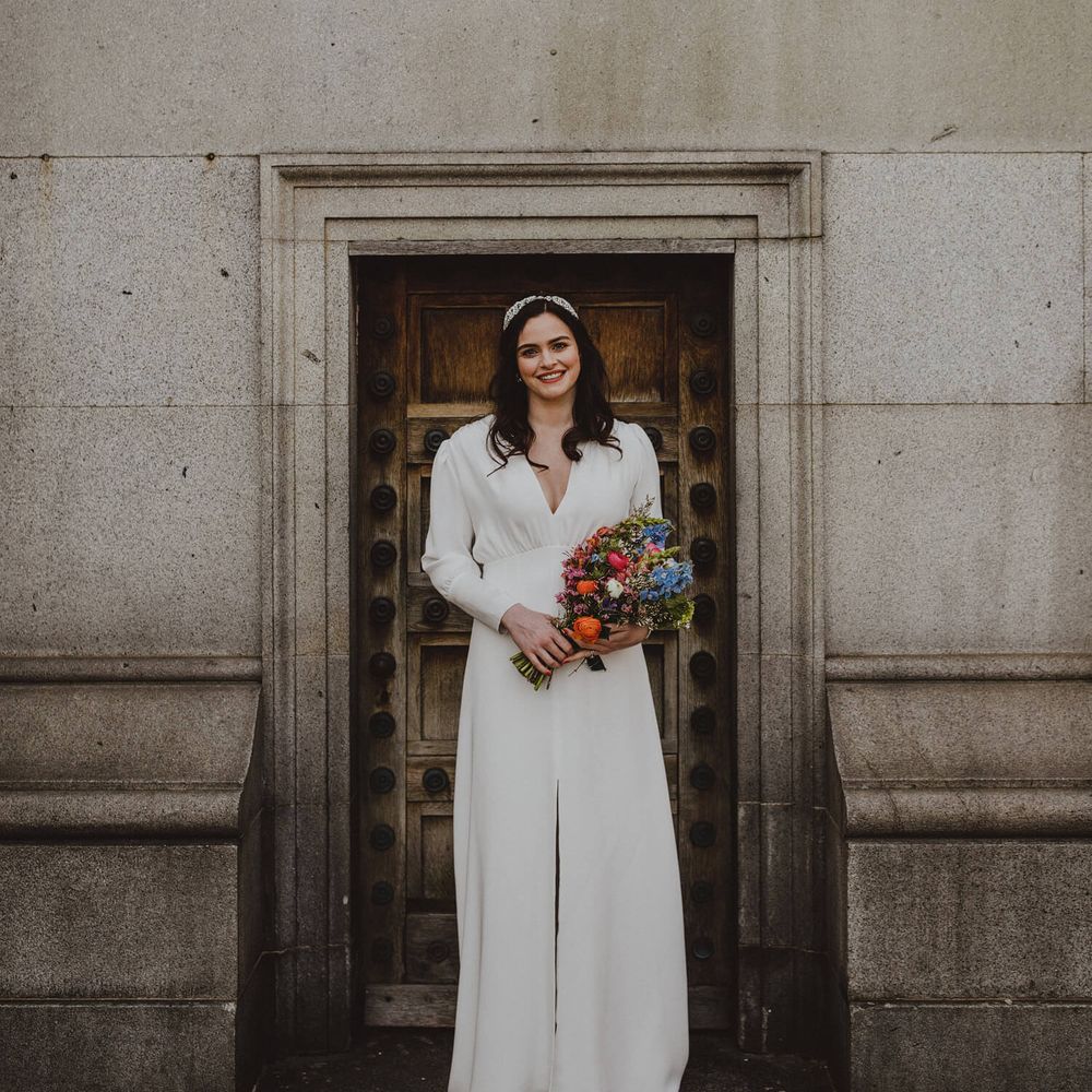 Bride outside London wedding venue wearing silk bridal gown with long sleeves and front slit, holding bright poppy wedding bouquet 