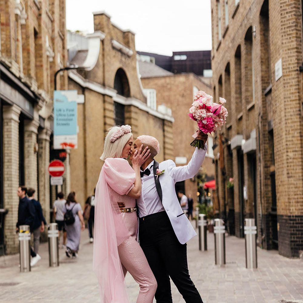 Bride in a pink jumpsuit kissing her bride in a white tuxedo jacket 