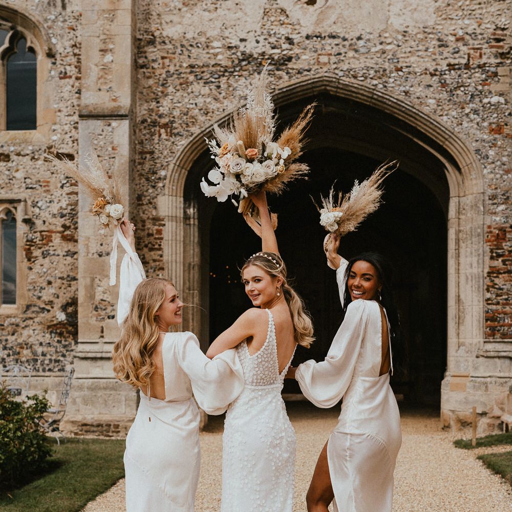 Bride in an appliqué wedding dress holding her dried flower bouquet in the air stood with her bridesmaids in champagne satin dresses 