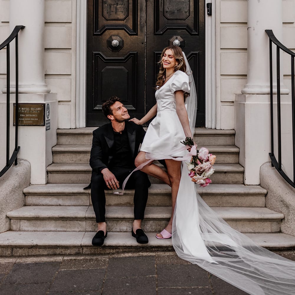 Bride in a short wedding dress with long wedding veil standing next to her husband dressed all in black sat on some steps in Mayfair 
