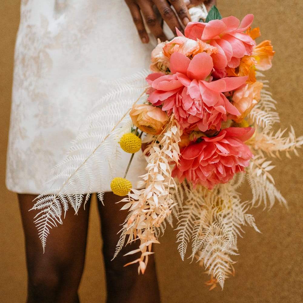 Bride wearing an embroidered mini skirt holding a bouquet of large pink dahlias, dried ferns and yellow craspedias