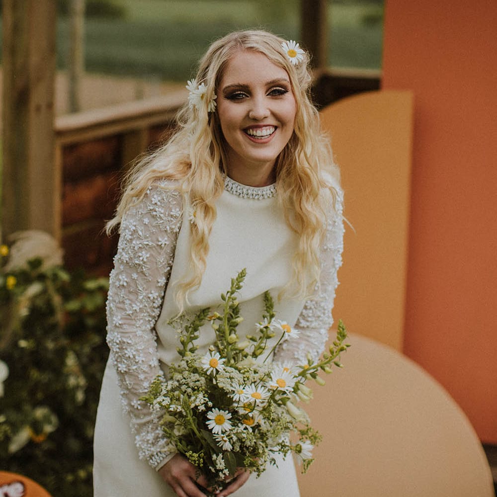 Sixties bride in a mini wedding dress with long wavy hair and wildflowers holding a daisy bouquet 