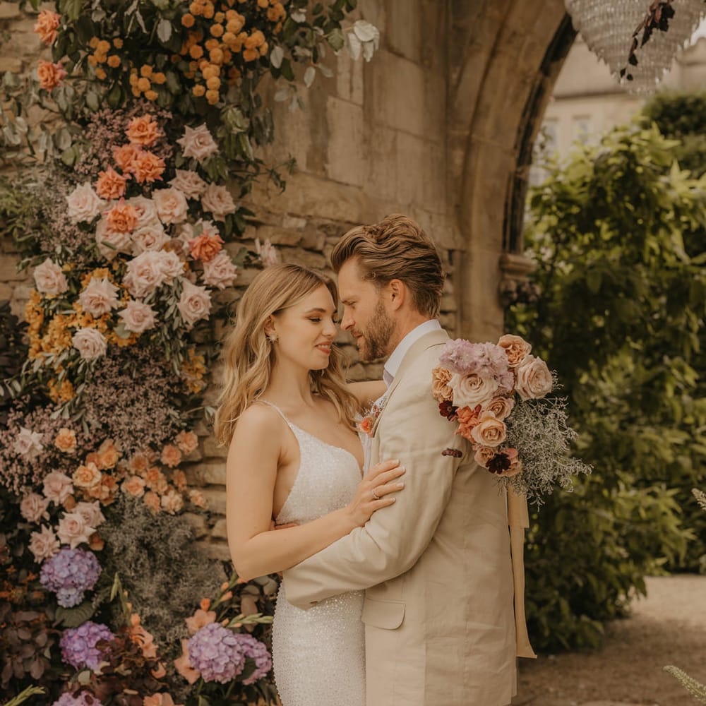 Bride with long wavy hair wearing a sparkly wedding dress embracing her groom in a beige suit with her pastel wedding bouquet over his shoulder 