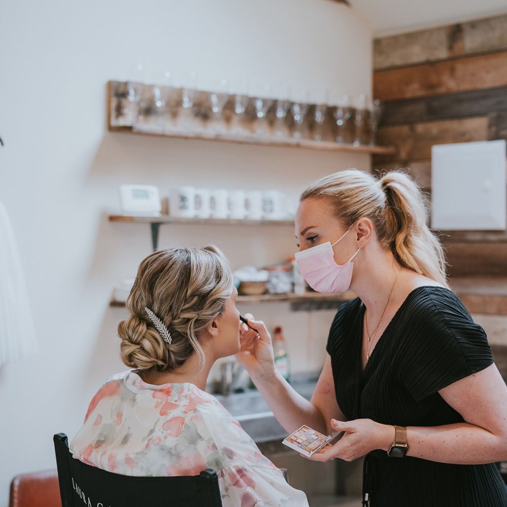 Bride with plaited bun and leaf hair pin sits having her make up done by woman in black jumpsuit and pink face mask before Tythe Barn wedding with barn wedding flowers