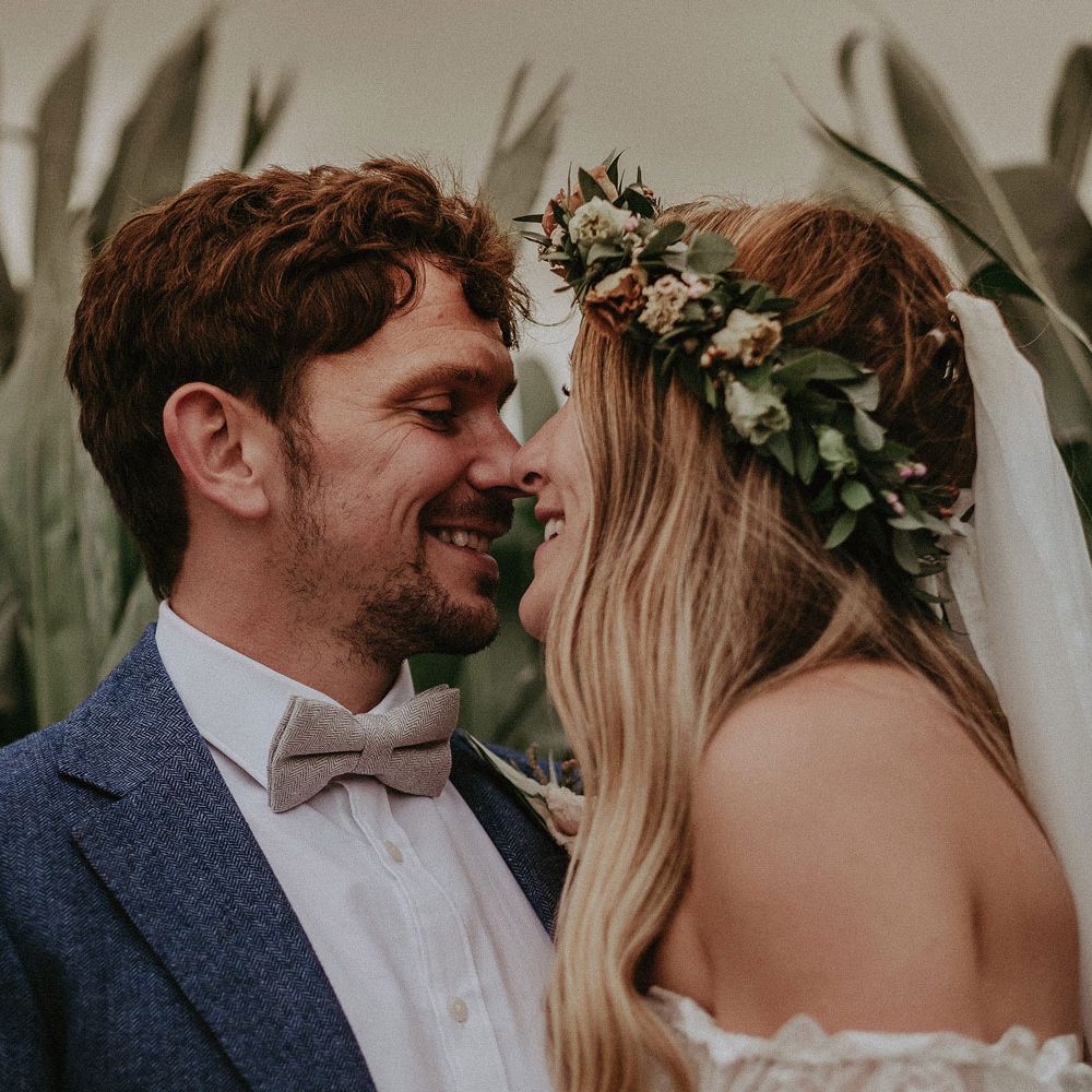 Bride in Grace Loves Lace wedding dress, veil and flower crown smiles with groom in blue suit jacket and sage green bow tie as they stand in corn field at Isle of Wight wedding with macrame wedding decor