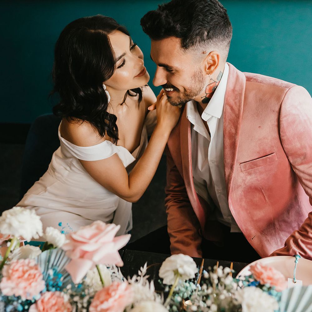 Groom in a gold shoulder wedding dress leaning in to kiss her groom in a pink velvet dinner jacket 