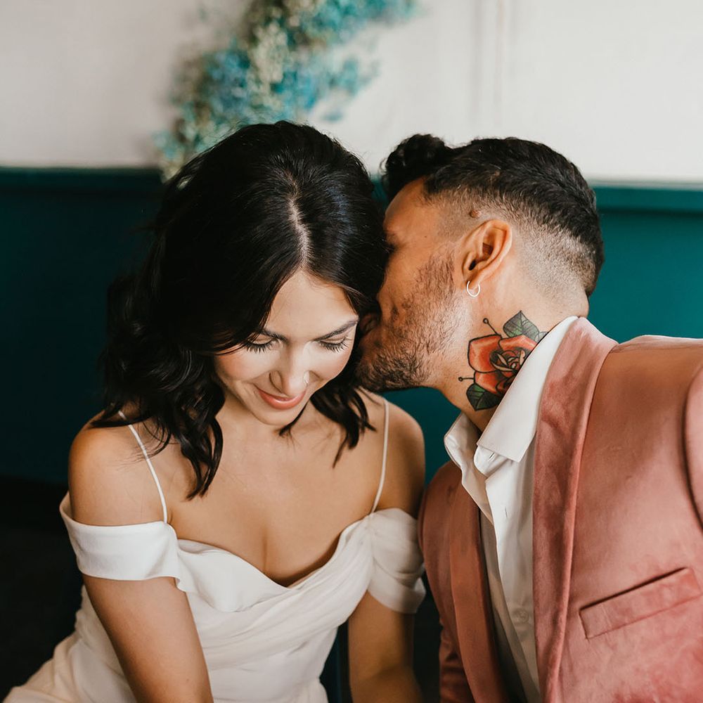 Groom in a pink velvet jacket with neck tattoo kissing his brides neck in a cold shoulder wedding dress 