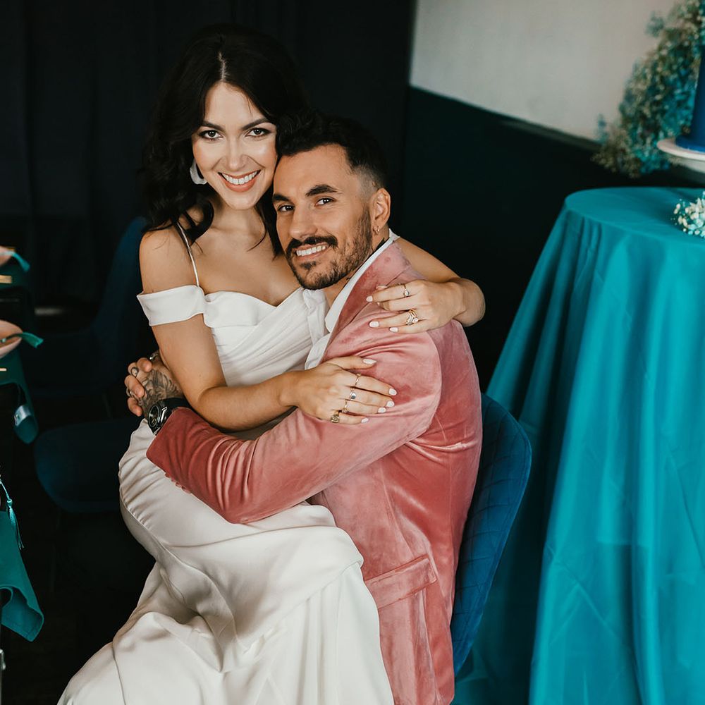 Bride in a cold shoulder wedding dress sitting on her grooms lap in a pink velvet jacket 