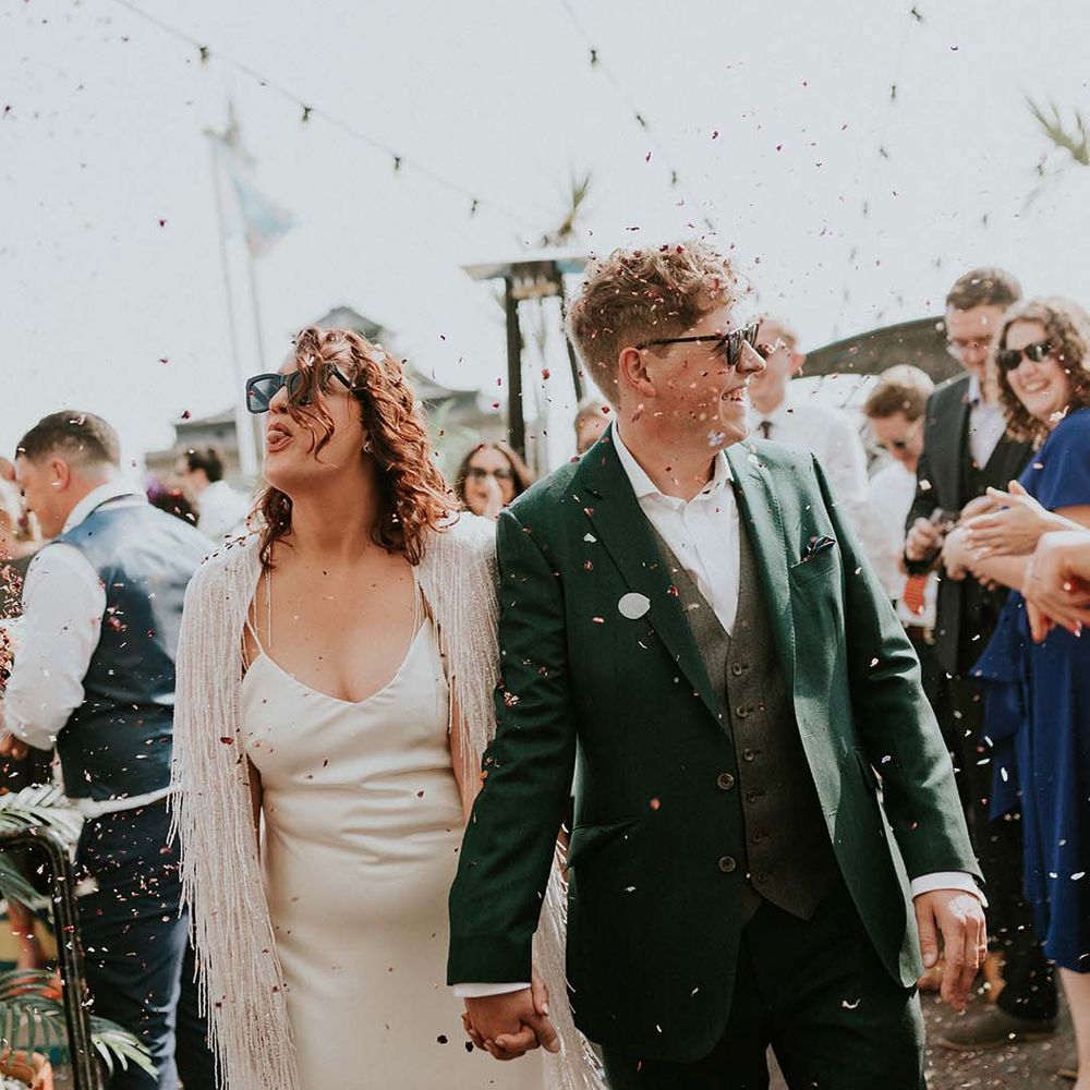 Bride & groom walk hand in hand through confetti as bride playfully sticks her tongue out to wedding guest
