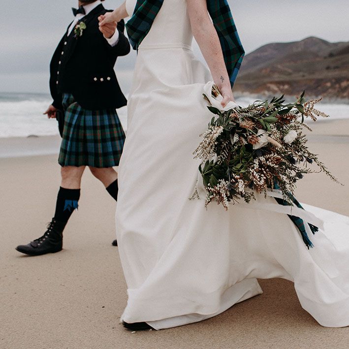 Bride holding her boho bouquet with anemones, wildflowers and foliage by her side as she walks along the beach 