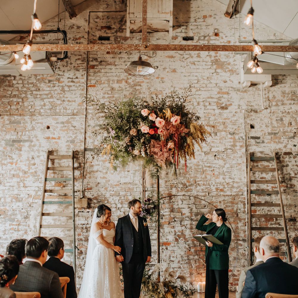 Bride in lace wedding dress and groom in blue check suit stand under mixed pink, yellow and green flower cloud with foliage and florals during wedding ceremony at Scale in Liverpool
