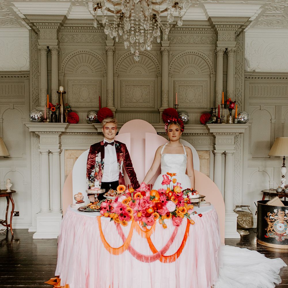 Two brides in a Burgundy velvet blazer and tulle skirt wedding dress holding hands in front of a  pink geometric backdrop, red, pink and orange flower arrangement and food by Baba Ganoush 