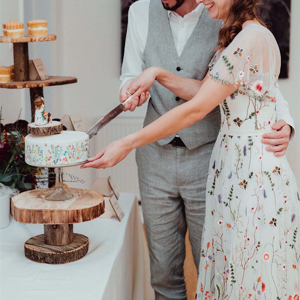 Bride in embroidered floral wedding dress with groom in grey waistcoat cut their white flowery wedding cake on tree trunk stand