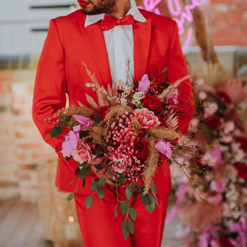 Groom in a red suit and bow tie holding a pink and red wedding bouquet at rustic barn wedding 