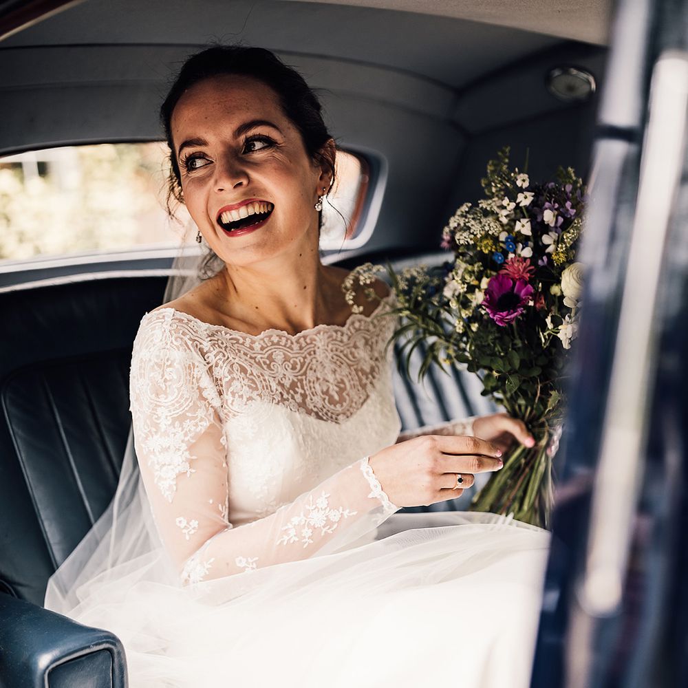 Bride smiles with diamond earrings in lace and satin wedding dress holding colourful wedding bouquet 