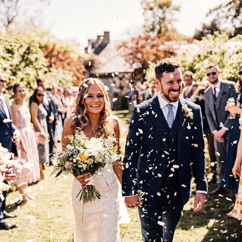 Bride carrying yellow bouquet holding hands with groom in blue suit have white confetti exit from ceremony 