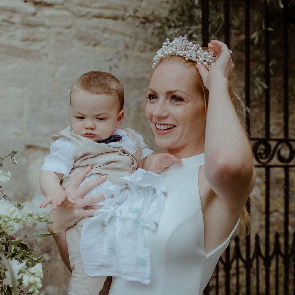 Bride adjusts her pearl headpiece as she holds a baby in her arms wearing beige trousers and waistcoat and a cute bow tie 