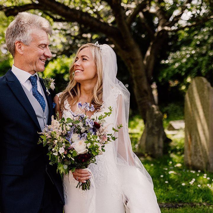 Bride in lace wedding dress with spring bouquet smiles at her father who wears a black suit with polka dot blue tie and handkerchief 
