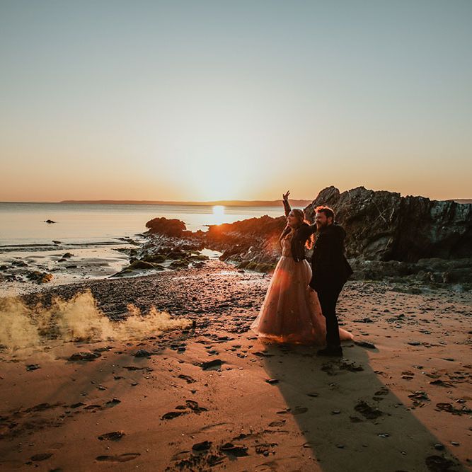 Bride & groom walk along beach during golden hour surrounded by colourful smoke bombs 