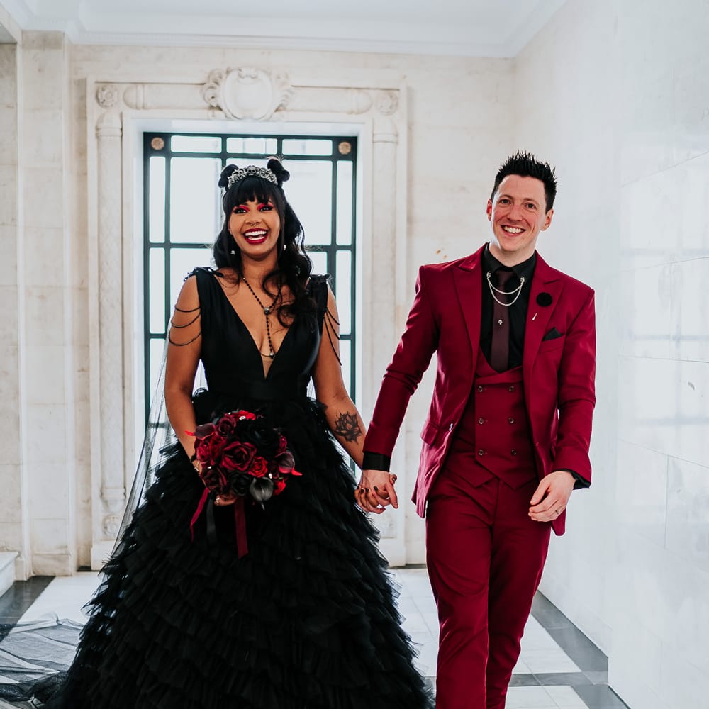 Bride wears black ruffled wedding dress with V shaped neckline and chain detailing to shoulders as she walks with her groom in wine red suit 